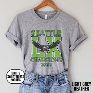 K&ouml;nnte beinhalten: Hellgraues meliertes T-Shirt mit Grafikdesign. Das Design zeigt den Text "SEATTLE LX CHAMPIONS 2026" in Gr&uuml;n und Schwarz, mit einer Adlerillustration. Das T-Shirt h&auml;ngt an einem Holzkleiderb&uuml;gel.