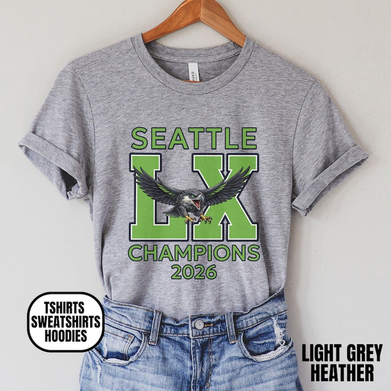 K&ouml;nnte beinhalten: Hellgraues meliertes T-Shirt mit Grafikdesign. Das Design zeigt den Text "SEATTLE LX CHAMPIONS 2026" in Gr&uuml;n und Schwarz, mit einer Adlerillustration. Das T-Shirt h&auml;ngt an einem Holzkleiderb&uuml;gel.