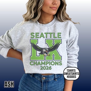 K&ouml;nnte beinhalten: Hellgraues Sweatshirt mit dem Text "SEATTLE LX CHAMPIONS 2026" in Gr&uuml;n und Wei&szlig;. Eine detaillierte Grafik eines Vogels mit ausgebreiteten Fl&uuml;geln ist mittig auf der Vorderseite. Das Sweatshirt hat auch den Text "TSHIRTS SWEATSHIRTS HOODIES".