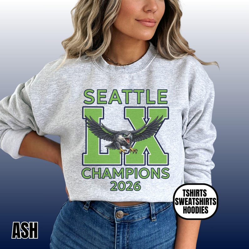 K&ouml;nnte beinhalten: Hellgraues Sweatshirt mit dem Text "SEATTLE LX CHAMPIONS 2026" in Gr&uuml;n und Wei&szlig;. Eine detaillierte Grafik eines Vogels mit ausgebreiteten Fl&uuml;geln ist mittig auf der Vorderseite. Das Sweatshirt hat auch den Text "TSHIRTS SWEATSHIRTS HOODIES".