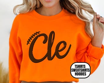 May include: Orange sweatshirt with the word "Cle" in a brown, stylized font. The letter "C" is decorated with a football design. The sweatshirt has a crew neck and long sleeves. The bottom right corner of the sweatshirt has a white circle with the words "Tshirts Sweatshirts Hoodies".