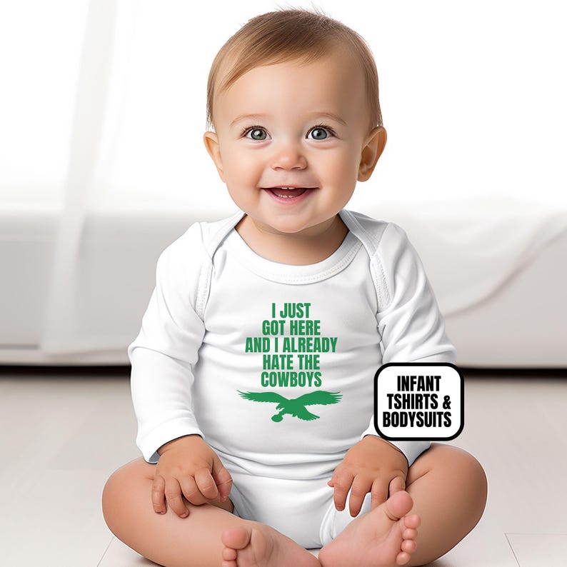 Puede incluir: Un body de manga larga para beb&eacute; en color blanco con texto verde que dice I JUST GOT HERE AND I ALREADY HATE THE COWBOYS sobre un gr&aacute;fico de un &aacute;guila verde. La prenda tiene un cuello redondo y mangas largas que cubren el torso superior.
