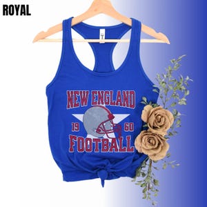 Pu&ograve; includere: Una canotta racerback blu reale con una stampa grafica che riporta il testo NEW ENGLAND FOOTBALL, i numeri 19 e 60, una stella bianca e un casco da football americano grigio con maschera rossa. L'orlo &egrave; annodato al centro sul davanti.