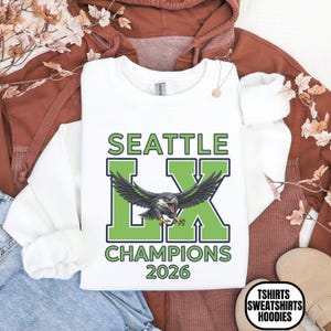 K&ouml;nnte beinhalten: Wei&szlig;es Sweatshirt mit dem Text "SEATTLE LX CHAMPIONS 2026" in Gr&uuml;n und Schwarz. Das Design zeigt einen Vogel mit ausgebreiteten Fl&uuml;geln. Teil einer Kollektion von T-Shirts, Sweatshirts und Hoodies.