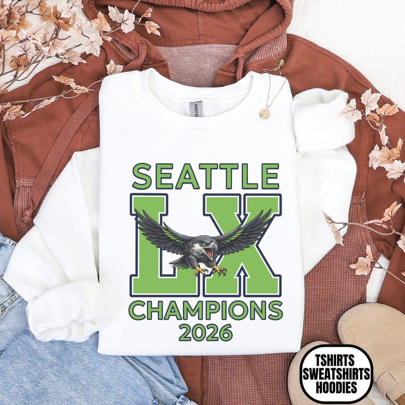 K&ouml;nnte beinhalten: Wei&szlig;es Sweatshirt mit dem Text "SEATTLE LX CHAMPIONS 2026" in Gr&uuml;n und Schwarz. Das Design zeigt einen Vogel mit ausgebreiteten Fl&uuml;geln. Teil einer Kollektion von T-Shirts, Sweatshirts und Hoodies.