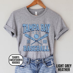 May include: Light grey heather t-shirt with "TAMPA BAY BASEBALL" in light blue. The design includes crossed baseball bats, a baseball, and the numbers "19" and "98". The shirt is hanging on a wooden hanger.