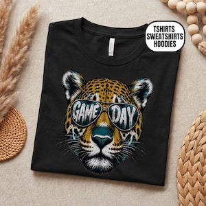 May include: A black t-shirt featuring a graphic of a leopard face wearing sunglasses. The lenses of the sunglasses display the text GAME DAY. A white circular label in the top right corner contains the text TSHIRTS SWEATSHIRTS HOODIES.