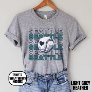 May include: Light grey heather t-shirt with the word "SEATTLE" stacked vertically and horizontally in white and teal. A baseball with a heart is in the center. The text "TSHIRTS SWEATSHIRTS HOODIES" is in a white circle.
