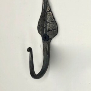 May include: A black metal leaf-shaped hook with a decorative design. The hook is attached to a white wall with a screw.