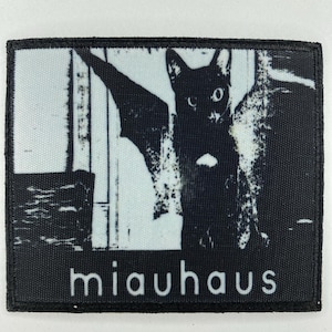 May include: Black and white patch with a cat-bat hybrid design. The image shows a cat with bat wings in front of a window. The word "miauhaus" is printed in white at the bottom.