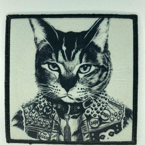 Puede incluir: Parche en blanco y negro con un gato con una chaqueta de cuero con tachuelas. El gato tiene una expresión seria y está centrado en un parche cuadrado con un borde negro. La chaqueta tiene parches que dicen "The Clash" y "Cash".