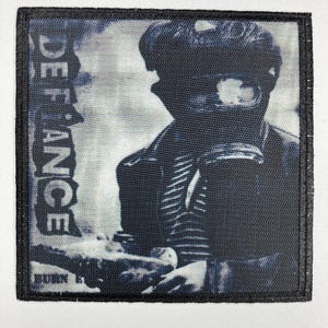 Op de afbeelding: Zwart-witte patch met het woord "Defiance" verticaal in wit gedrukt. De patch toont een persoon met een zwart gasmasker en een gestreept shirt. De tekst "Burn" is onderaan de patch in wit gedrukt.