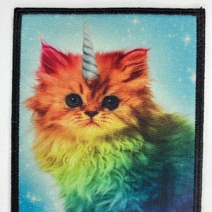 Puede incluir: Un gato de colores arcoíris con un cuerno de unicornio plateado en la frente. El gato mira directamente al espectador sobre un fondo azul con estrellas blancas.