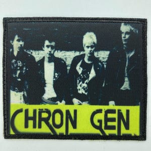Op de afbeelding: Een rechthoekige patch met een zwarte rand, met een zwart-wit afbeelding van vier personen. De patch heeft een gele banner aan de onderkant met de tekst "CHRON GEN" in zwarte, opvallende letters.