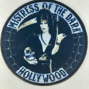Puede incluir: Parche redondo con una mujer de pelo oscuro y maquillaje, sosteniendo una calavera y una guadaña. El parche tiene las palabras "Mistress of the Dark" y "Hollywood" en un diseño circular, con un patrón de telaraña alrededor del borde.