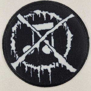 Puede incluir: Un parche redondo negro con un diseño blanco estilizado. El diseño presenta una forma de calavera con acentos goteantes y elementos tachados. El parche tiene un borde cosido.