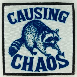 May include: Square patch with a white background, a blue raccoon illustration, and the words "CAUSING CHAOS" in blue. The patch has a black border.