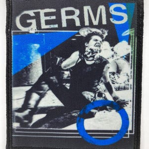 May include: A square fabric patch with a black border featuring a black and white photograph of a person performing on stage. The word GERMS is printed in white capital letters at the top. A bright blue circle and blue geometric shapes are visible.