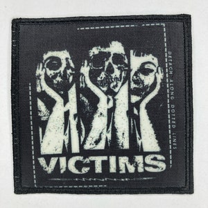 Pode incluir: Patch preto e branco com um gráfico de três figuras semelhantes a caveiras, com as mãos cobrindo as orelhas. A palavra "VICTIMS" é impressa em letras brancas em negrito abaixo das figuras. A frase "DETACH ALONG DOTTED LINES" é impressa verticalmente no lado direito.