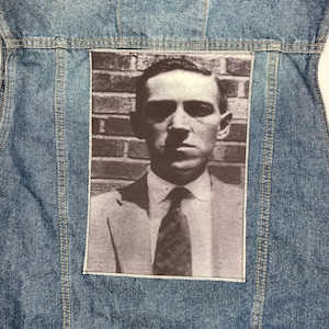 Op de afbeelding: Een blauwe denim jas met een gestikt patch waarop een zwart-witfoto van een man in pak en stropdas te zien is. De man kijkt recht in de camera met een serieuze uitdrukking.