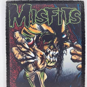 May include: A rectangular fabric patch with a black stitched border featuring a graphic illustration of a skeletal figure with a prominent eye and sharp teeth. The text MISFITS is printed in a stylized green font at the top against a dark background.