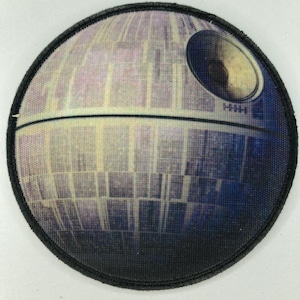 Op de afbeelding: Ronde patch met een gedetailleerde afbeelding van de Death Star uit Star Wars. Het ontwerp toont een grote ronde structuur met een opvallende schotel en ingewikkelde oppervlakte details. De patch heeft een zwarte rand.