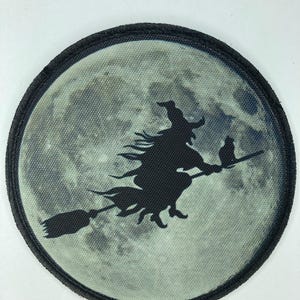 Könnte beinhalten: Runder Aufnäher mit einer schwarzen Silhouette einer Hexe auf einem Besen vor einem Vollmond. Der Mond ist hellgrau und der Aufnäher hat einen schwarzen Rand. Eine Katze reitet mit der Hexe. Halloween-Thema.