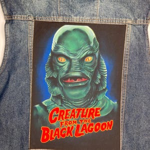 Pode incluir: Um remendo preto e verde com uma criatura da lagoa negra. A criatura tem um rosto verde com olhos grandes e uma boca larga. O texto "Creature from the Black Lagoon" está em letras vermelhas.