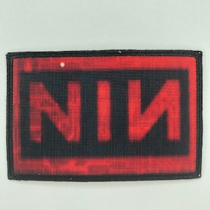 Puede incluir: Un parche rectangular bordado con un borde negro y un fondo rojo de textura desgastada. Las letras NIИ están impresas en una fuente negra, negrita y sin remates, con la última N orientada en forma de espejo.