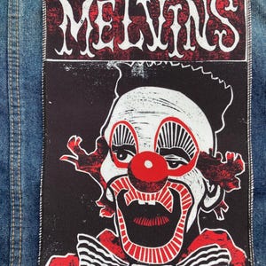 May include: A rectangular fabric patch featuring a graphic of a clown with white face paint, a red nose, and red and black details. The word MELVINS is printed in white stylized lettering at the top against a red and black background.