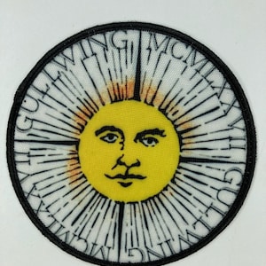 May include: Circular patch featuring a yellow sun with a face, surrounded by white rays and a black border. The sun has a mustache and is centered on the patch. The text "GUT WING MCMXCI" is written around the edge of the patch.