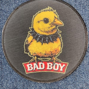 Peut inclure: Un écusson brodé circulaire représentant un poussin jaune portant un collier à pointes noir. Sous le poussin, une bannière rouge à bordure blanche contient le texte BAD BOY en lettres capitales blanches. L'écusson a une bordure noire.
