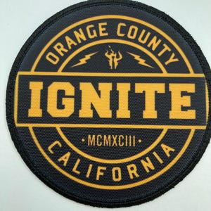 May include: A circular embroidered patch featuring a black background with yellow text and graphics. The text reads ORANGE COUNTY, IGNITE, and CALIFORNIA, with the Roman numeral MCMXCIII in the centre, accompanied by flame and lightning bolt icons.