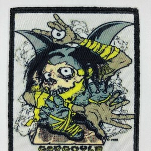 Könnte beinhalten: Quadratischer Aufnäher mit einem Cartoon-Gargoyle-Design. Der Gargoyle hat ein schädelähnliches Gesicht, fledermausartige Ohren und ist in gelbe Bandagen gewickelt. Der Aufnäher hat einen schwarzen Rand und das Wort "GARGOYLE" ist unten aufgedruckt.