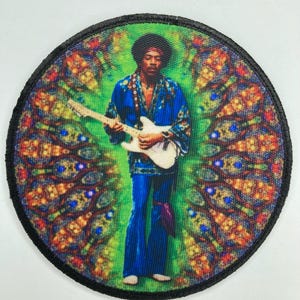 Puede incluir: Un parche bordado circular que presenta un gráfico colorido de un músico con peinado afro tocando una guitarra eléctrica blanca. La figura viste una camisa azul estampada y pantalones azules sobre un fondo vibrante de color verde y naranja.
