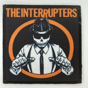 May include: A square fabric patch featuring a black background with an orange circle. The text THE INTERRUPTERS appears in orange at the top. A central graphic shows a skeleton wearing a fedora, tie, and suspenders, with the words TRICK and TREAT on its hands.