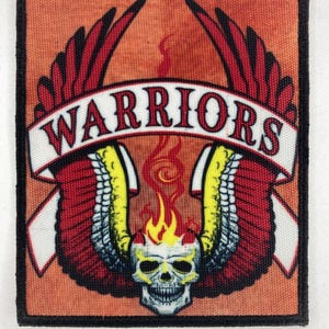 Puede incluir: Parche cuadrado con un diseño de calavera, alas y llamas. El parche presenta una calavera con llamas que se elevan por encima, flanqueada por alas rojas y amarillas. La palabra "WARRIORS" se muestra en una pancarta blanca. El parche tiene un borde negro.
