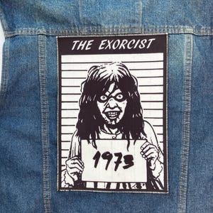 Pode incluir: Um remendo preto e branco com uma fotografia policial de um demônio do filme "O Exorcista" com o ano "1973" impresso em um cartaz. O remendo é costurado em uma jaqueta jeans azul.