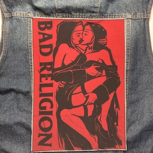 Puede incluir: Chaqueta vaquera azul con un parche rojo. El parche muestra una ilustración negra de dos figuras abrazándose, con las palabras "BAD RELIGION" impresas verticalmente. La imagen representa una escena de besos.