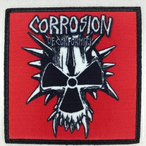 Op de afbeelding: Een vierkante patch met een rode achtergrond en een zwarte rand. De patch heeft een zwart-wit schedelontwerp met spikes en de woorden "CORROSION OF CONFORMITY" in witte, gestileerde letters.