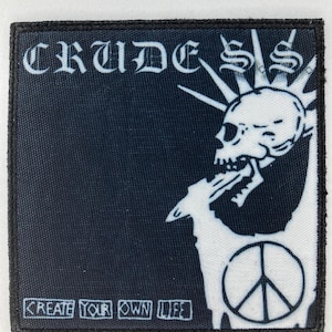 Puede incluir: Parche en blanco y negro con la palabra "CRUDE SS" en fuente gótica. Una calavera blanca con cresta mohawk y símbolo de la paz está a la derecha. El texto "CREATE YOUR OWN LIFE" está en la parte inferior.