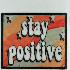 Op de afbeelding: Een rechthoekige stoffen patch met een zwarte rand met de tekst stay positive in witte bubbelletters met zwarte omlijning. De achtergrond bestaat uit golvende oranje, perzikkleurige en lichtgrijze strepen versierd met kleine witte sterren.