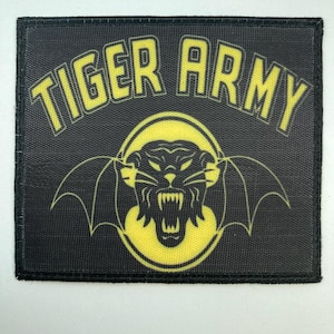 May include: A rectangular black fabric patch featuring the yellow text TIGER ARMY arched across the top. Below the text is a yellow graphic of a snarling panther head with bat-like wings, enclosed within a yellow oval border on a black background.