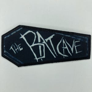 Puede incluir: Un parche bordado con forma de ataúd, borde negro y fondo azul oscuro. El texto "THE BAT CAVE" está bordado en el centro con una tipografía blanca, estilizada y dentada, que presenta un aspecto desgastado y envejecido.