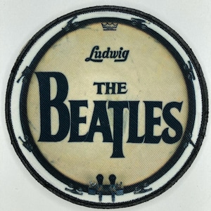 Può includere: Toppa circolare con la scritta "The Beatles" in blu navy. La toppa ha uno sfondo color crema con il logo "Ludwig" e una corona. Il bordo è nero con un contorno bianco.