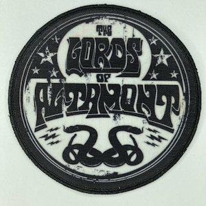 May include: A circular black and white embroidered patch featuring the text The Lords of Altamont in a stylized font. The design includes two intertwined snakes at the bottom and four small stars on the sides, all enclosed within a thick black border.