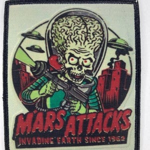 Könnte beinhalten: Quadratischer Aufnäher mit einem grünen Alien aus dem Film "Mars Attacks", der eine Strahlenkanone hält. Der Aufnäher trägt den Text "Mars Attacks" und "Invading Earth Since 1962". Das Alien hat ein großes Gehirn.