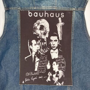 Pode incluir: Um colete de ganga azul com um grande patch preto com o nome da banda "bauhaus" em branco. O patch inclui uma caveira, uma cruz e ilustrações de pessoas. O texto no patch inclui "LIPS IN WSIMAR 150 YBARS BALIHAUS" e "SNARY FOR VAMPIRSA & UNDBAY".