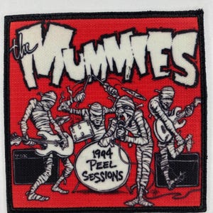 Puede incluir: Un parche rectangular con borde negro presenta un fondo rojo y el nombre de la banda "The Mummies" en blanco. La imagen representa cuatro figuras con aspecto de momia tocando instrumentos. El texto "1994 Peel Sessions" está en un tambor.