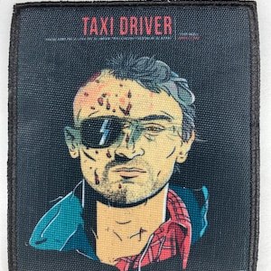 Peut inclure: Un patch brodé noir et blanc avec le texte "TAXI DRIVER" en lettres rouges. Le patch présente un gros plan sur le visage d'un homme avec du sang et un bandage sur le front. Il porte une veste bleue et une chemise à carreaux rouge et blanche.